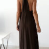 Robe longue marron dos nu Carla pour femme, coupe fluide et élégante, portée sur une plage en bord de mer sous le soleil d'été.