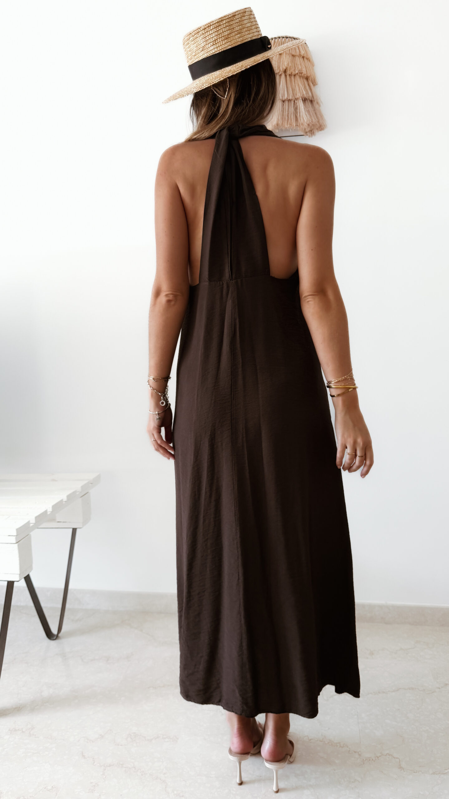 Robe longue marron dos nu Carla pour femme, coupe fluide et élégante, portée sur une plage en bord de mer sous le soleil d'été.