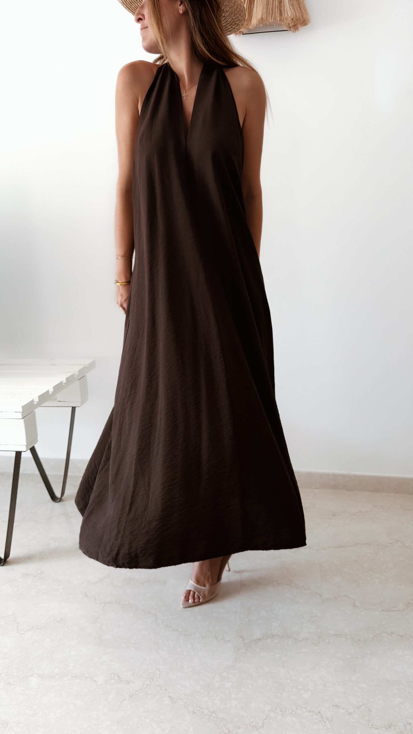 Robe longue marron dos nu Carla pour femme, coupe fluide et élégante, portée sur une plage en bord de mer sous le soleil d'été.