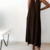 Robe longue marron dos nu Carla pour femme, coupe fluide et élégante, portée sur une plage en bord de mer sous le soleil d'été.