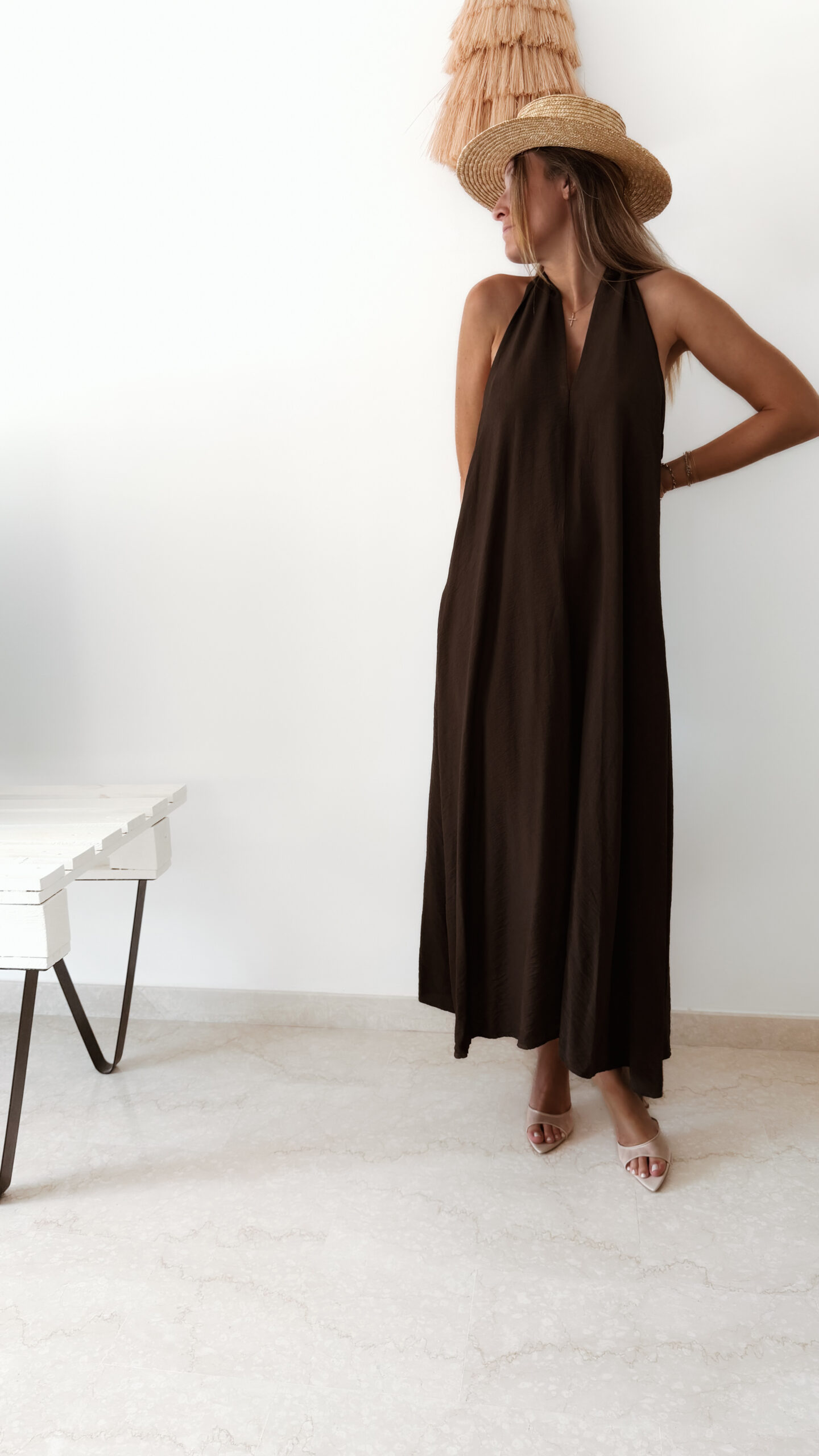 Robe longue marron dos nu Carla pour femme, coupe fluide et élégante, portée sur une plage en bord de mer sous le soleil d'été.