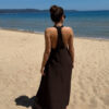 Robe longue marron dos nu Carla pour femme, coupe fluide et élégante, portée sur une plage en bord de mer sous le soleil d'été.