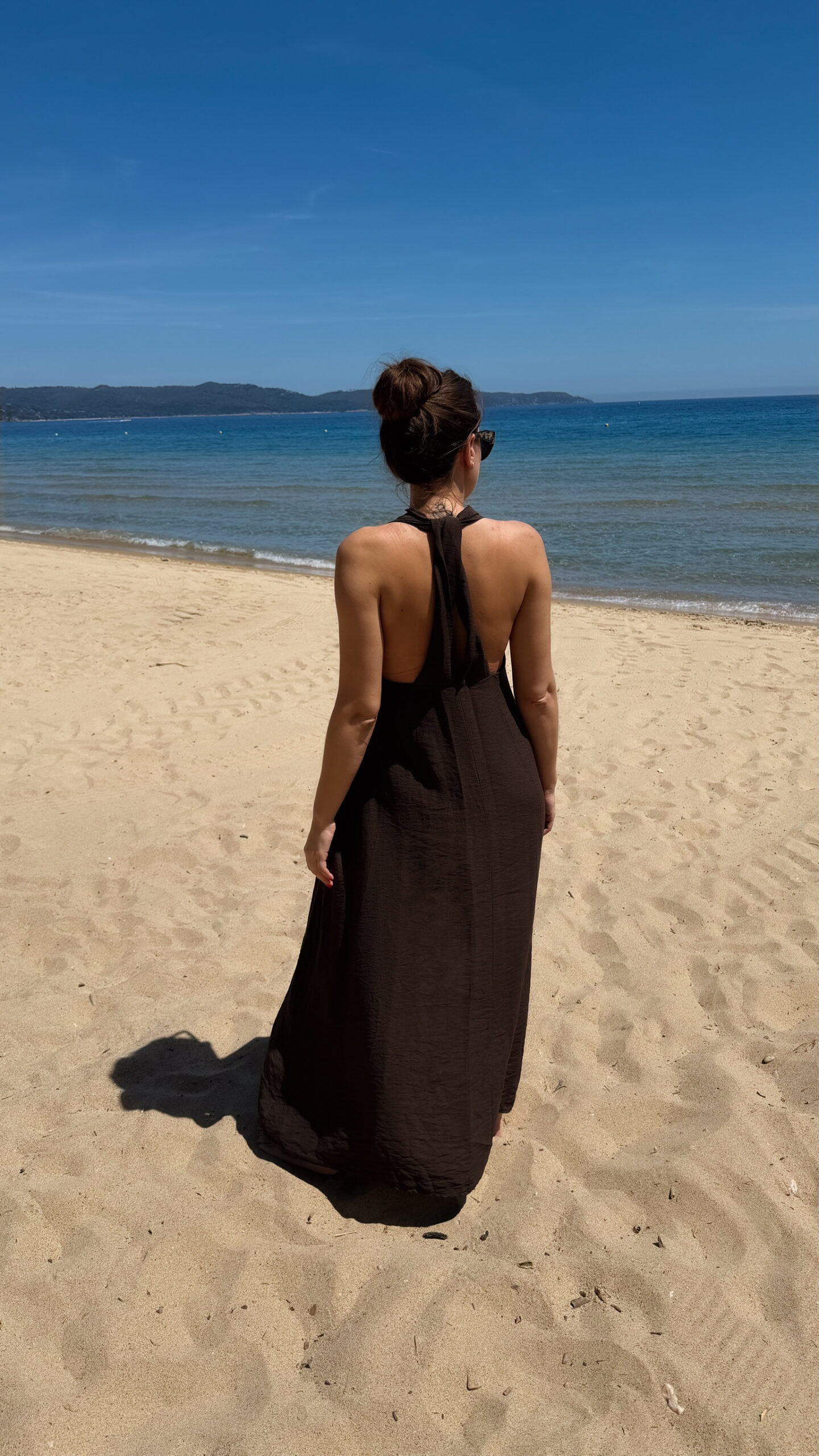 Robe longue marron dos nu Carla pour femme, coupe fluide et élégante, portée sur une plage en bord de mer sous le soleil d'été.