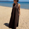 Robe longue marron dos nu Carla pour femme, coupe fluide et élégante, portée sur une plage en bord de mer sous le soleil d'été.