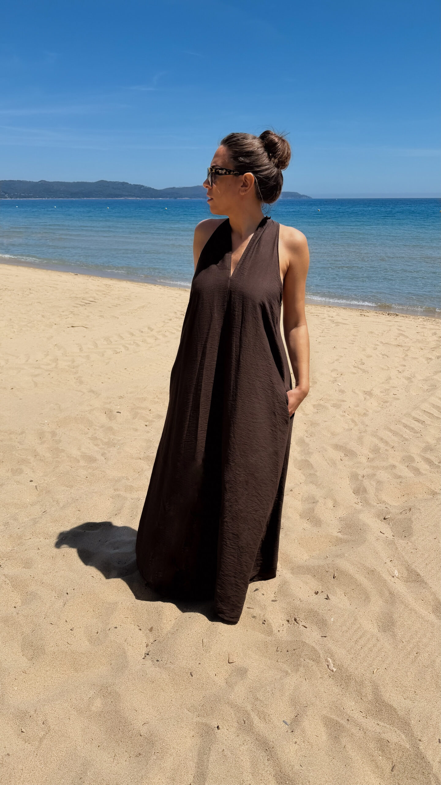Robe longue marron dos nu Carla pour femme, coupe fluide et élégante, portée sur une plage en bord de mer sous le soleil d'été.