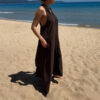 Robe longue marron dos nu Carla pour femme, coupe fluide et élégante, portée sur une plage en bord de mer sous le soleil d'été.