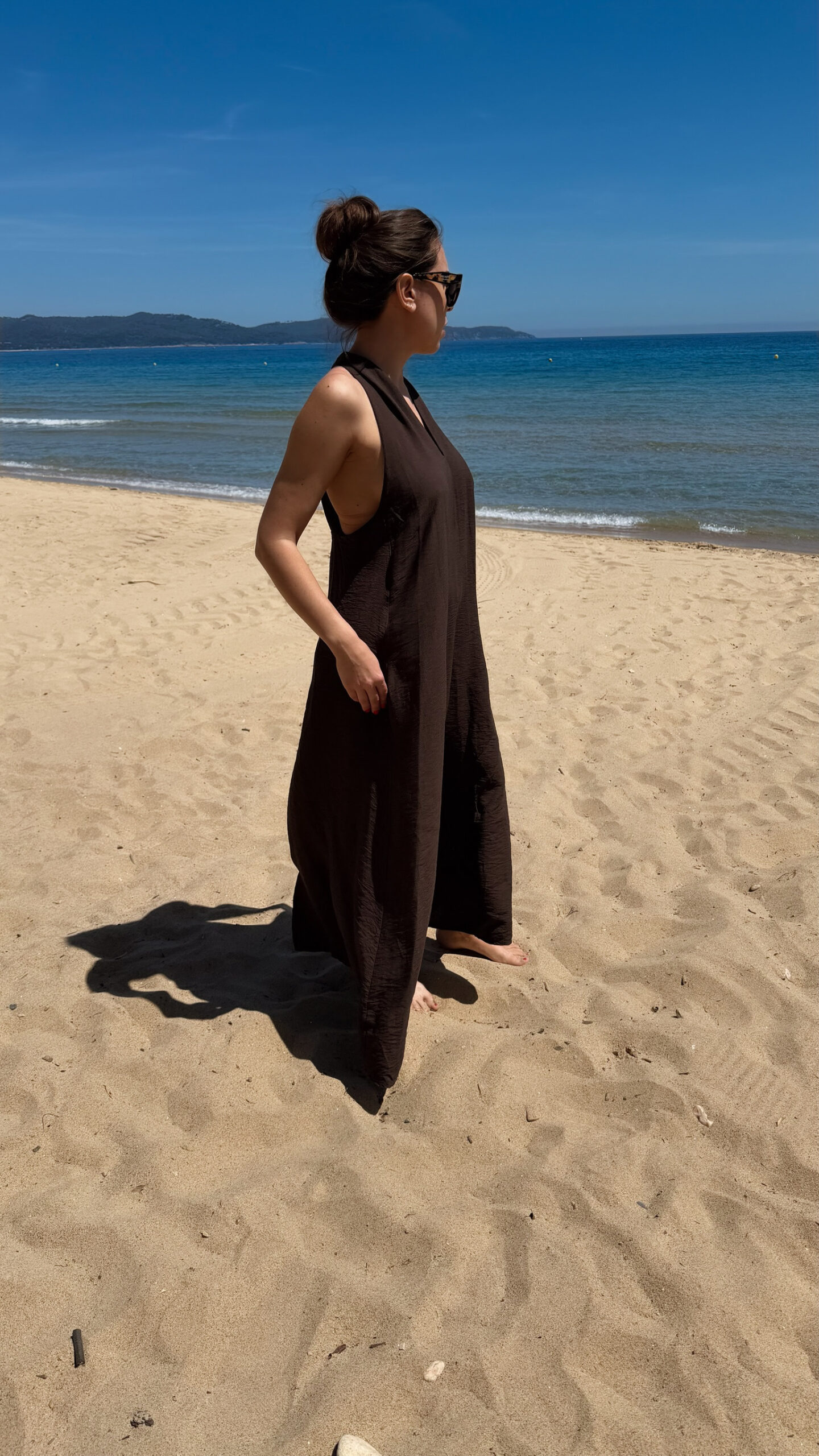 Robe longue marron dos nu Carla pour femme, coupe fluide et élégante, portée sur une plage en bord de mer sous le soleil d'été.
