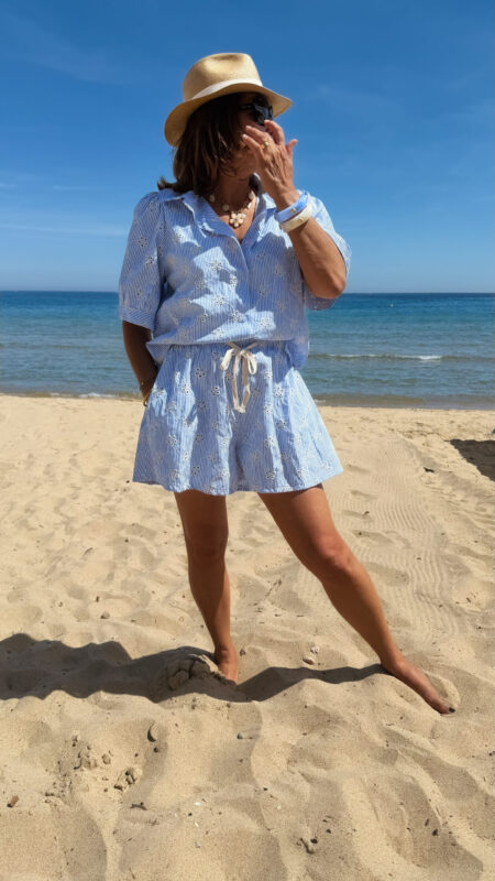 Ensemble Ninon femme bleu ciel et blanc brodé, composé d'une blouse légère et d'un short assorti, porté sur une plage en été.