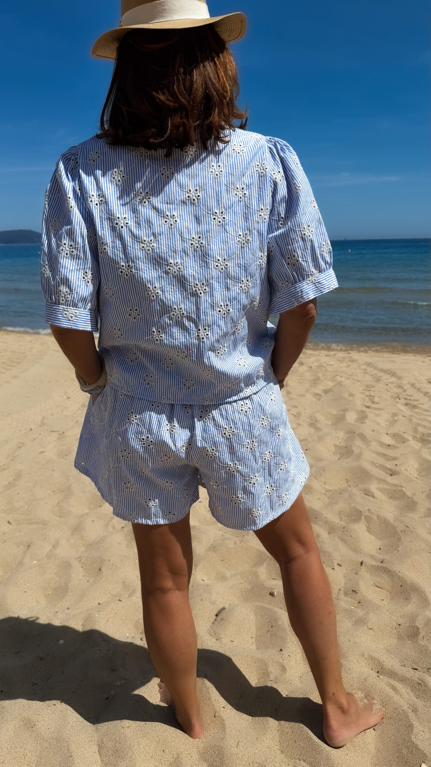 Ensemble Ninon femme bleu ciel et blanc brodé, composé d'une blouse légère et d'un short assorti, porté sur une plage en été.