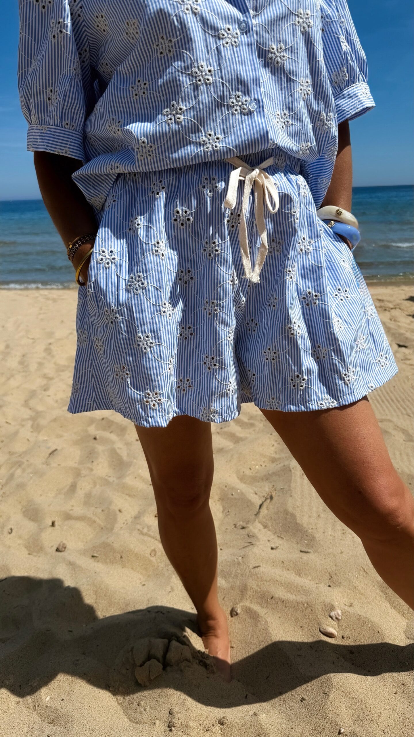 Ensemble Ninon femme bleu ciel et blanc brodé, composé d'une blouse légère et d'un short assorti, porté sur une plage en été.