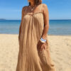 Femme portant une robe longue camel en gaze de coton sur une plage ensoleillée, avec chapeau et lunettes de soleil.