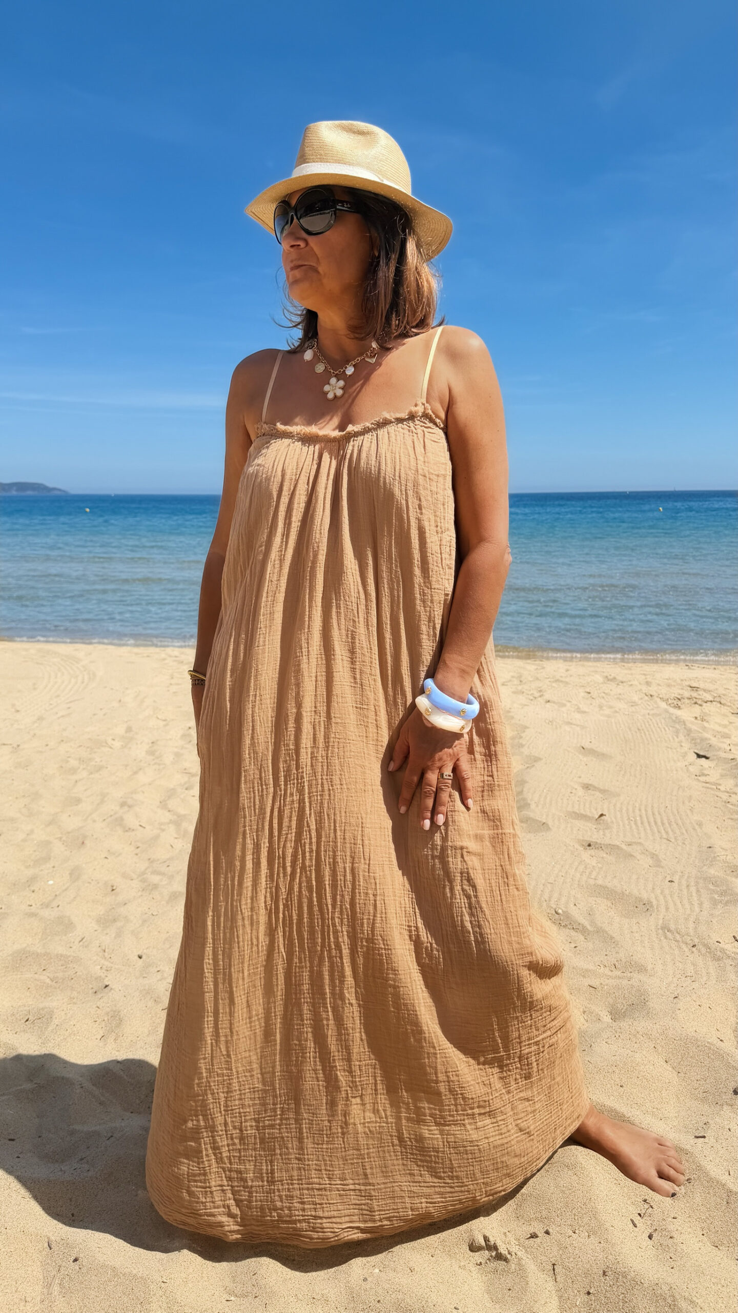 Femme portant une robe longue camel en gaze de coton sur une plage ensoleillée, avec chapeau et lunettes de soleil.