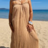 Femme portant une robe longue camel en gaze de coton sur une plage ensoleillée, avec chapeau et lunettes de soleil.