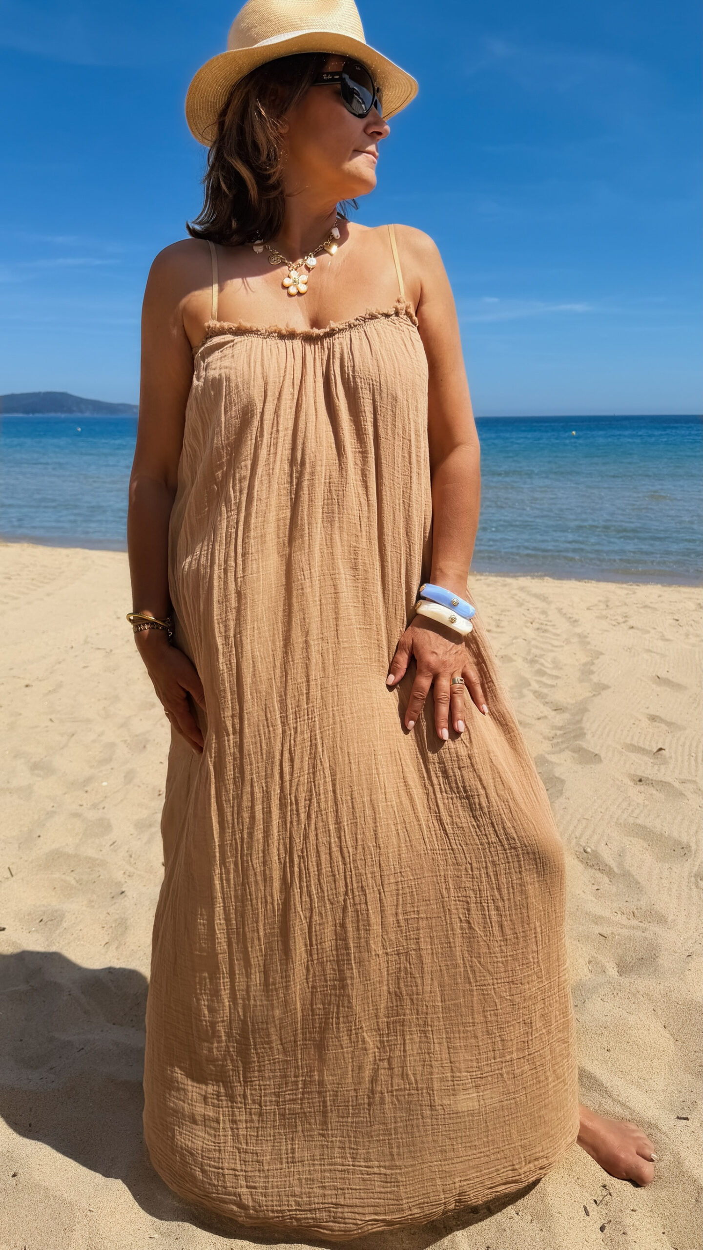 Femme portant une robe longue camel en gaze de coton sur une plage ensoleillée, avec chapeau et lunettes de soleil.