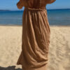 Femme portant une robe longue camel en gaze de coton sur une plage ensoleillée, avec chapeau et lunettes de soleil.