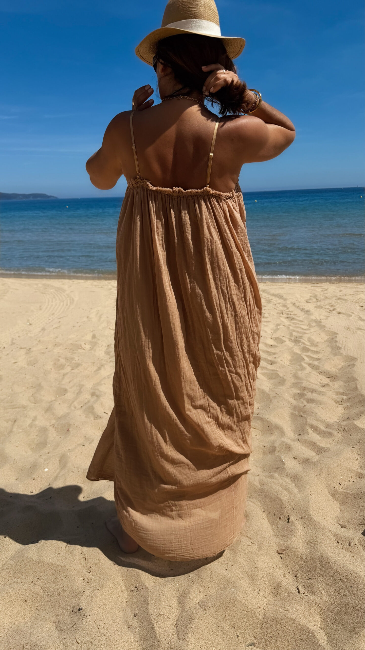 Femme portant une robe longue camel en gaze de coton sur une plage ensoleillée, avec chapeau et lunettes de soleil.