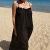 Femme portant une robe longue noire en gaze de coton sur une plage ensoleillée, avec chapeau et lunettes de soleil.