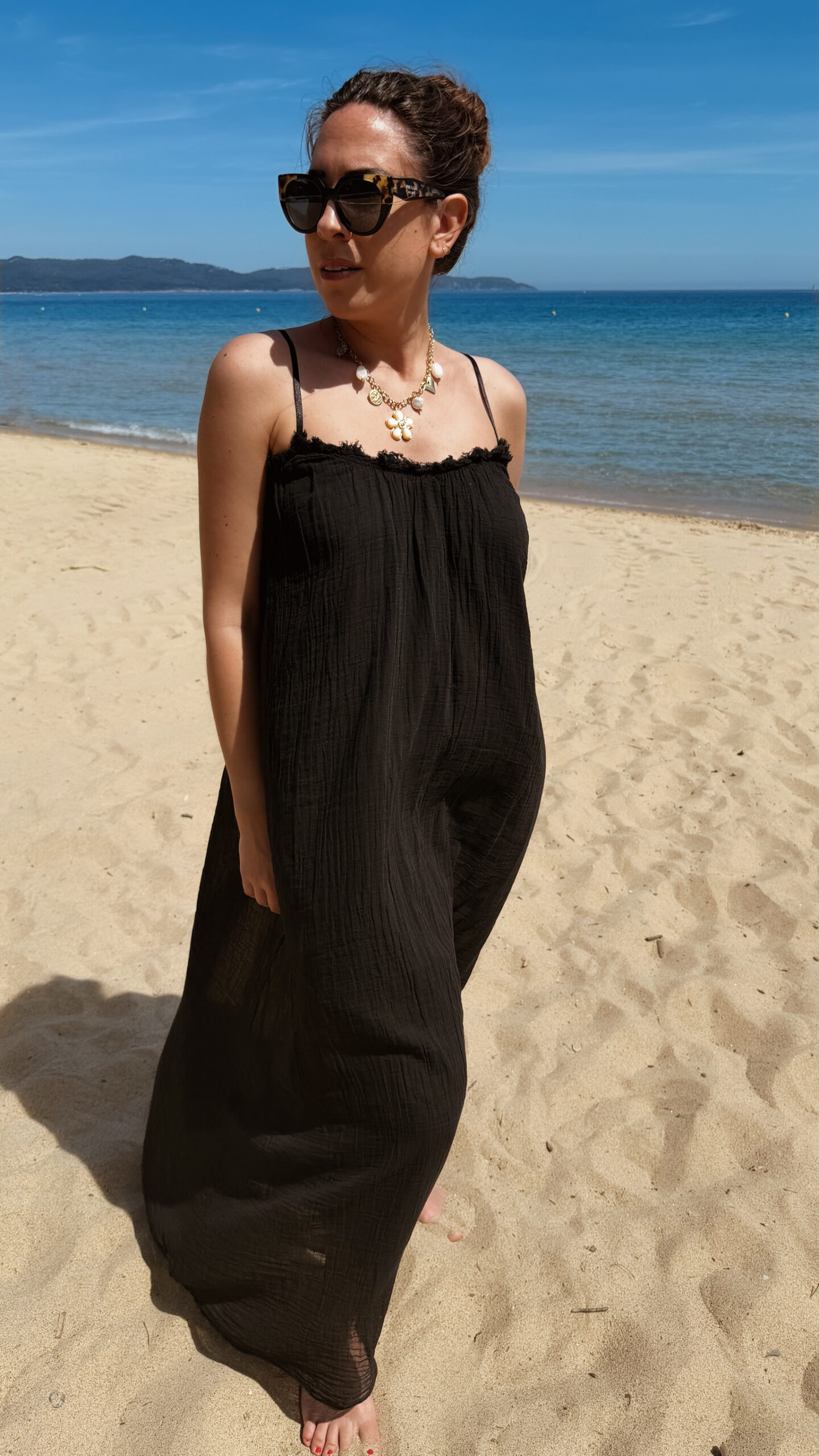 Femme portant une robe longue noire en gaze de coton sur une plage ensoleillée, avec chapeau et lunettes de soleil.