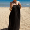 Femme portant une robe longue noire en gaze de coton sur une plage ensoleillée, avec chapeau et lunettes de soleil.