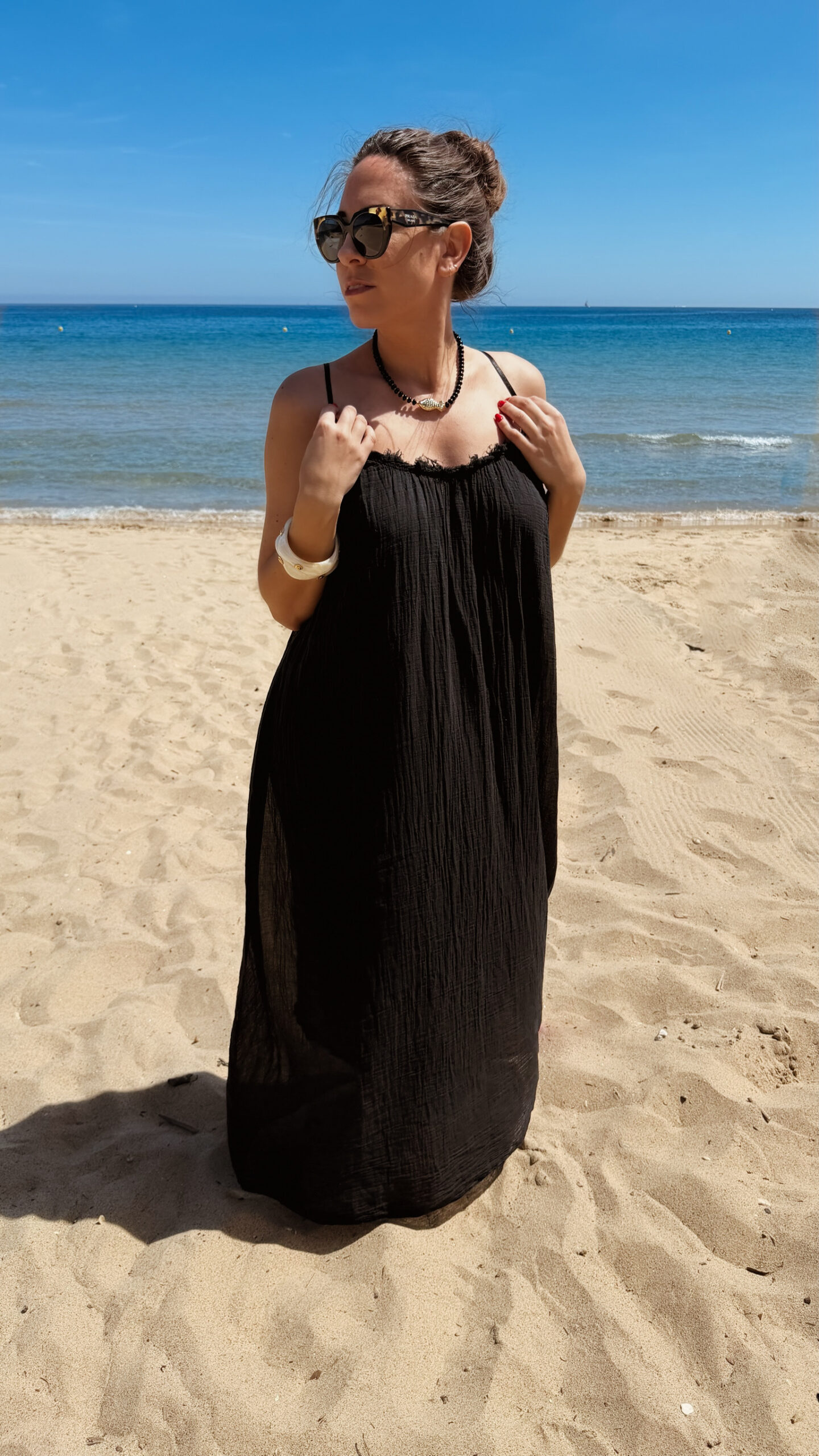 Femme portant une robe longue noire en gaze de coton sur une plage ensoleillée, avec chapeau et lunettes de soleil.