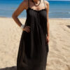 Femme portant une robe longue noire en gaze de coton sur une plage ensoleillée, avec chapeau et lunettes de soleil.