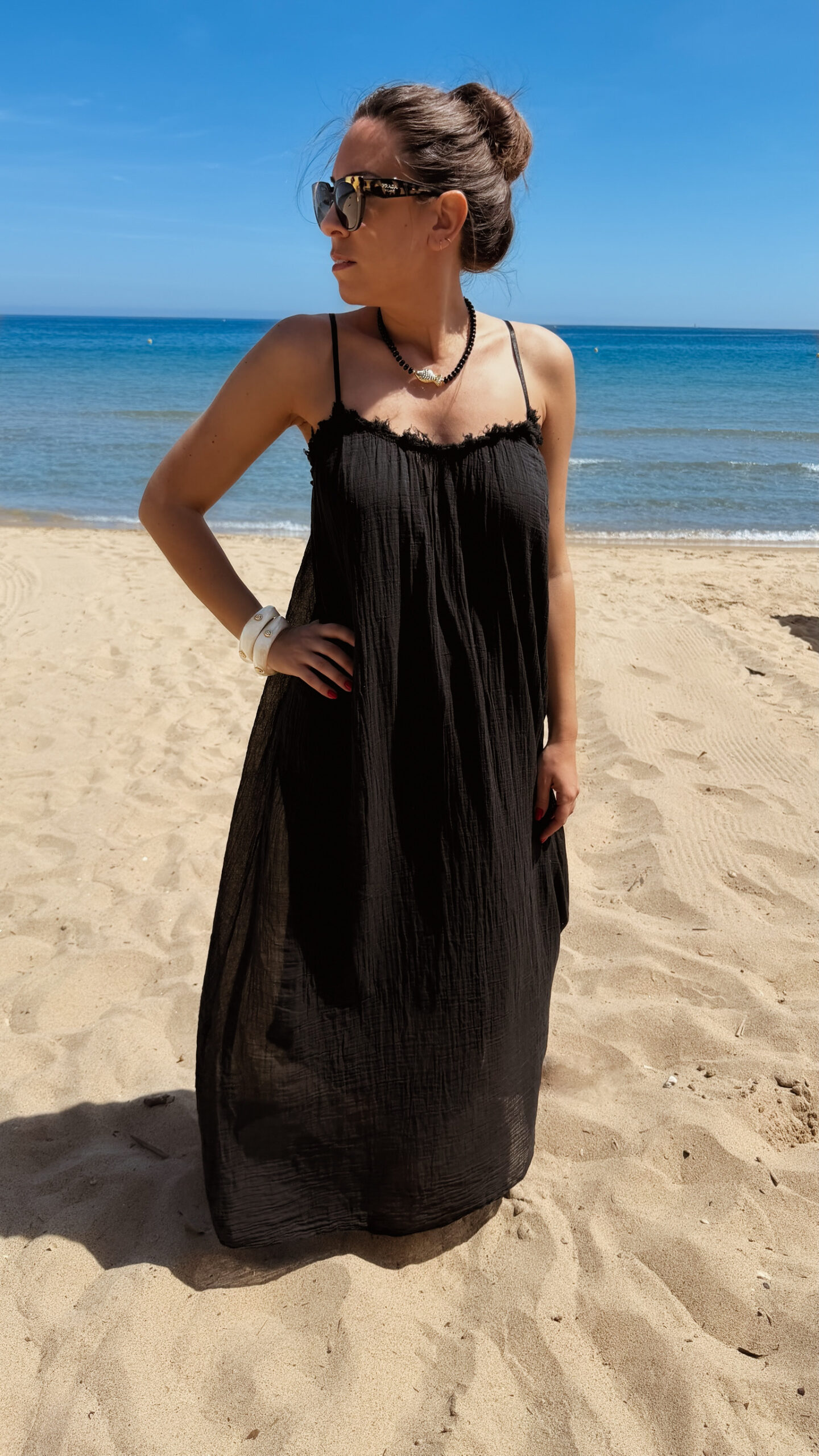 Femme portant une robe longue noire en gaze de coton sur une plage ensoleillée, avec chapeau et lunettes de soleil.