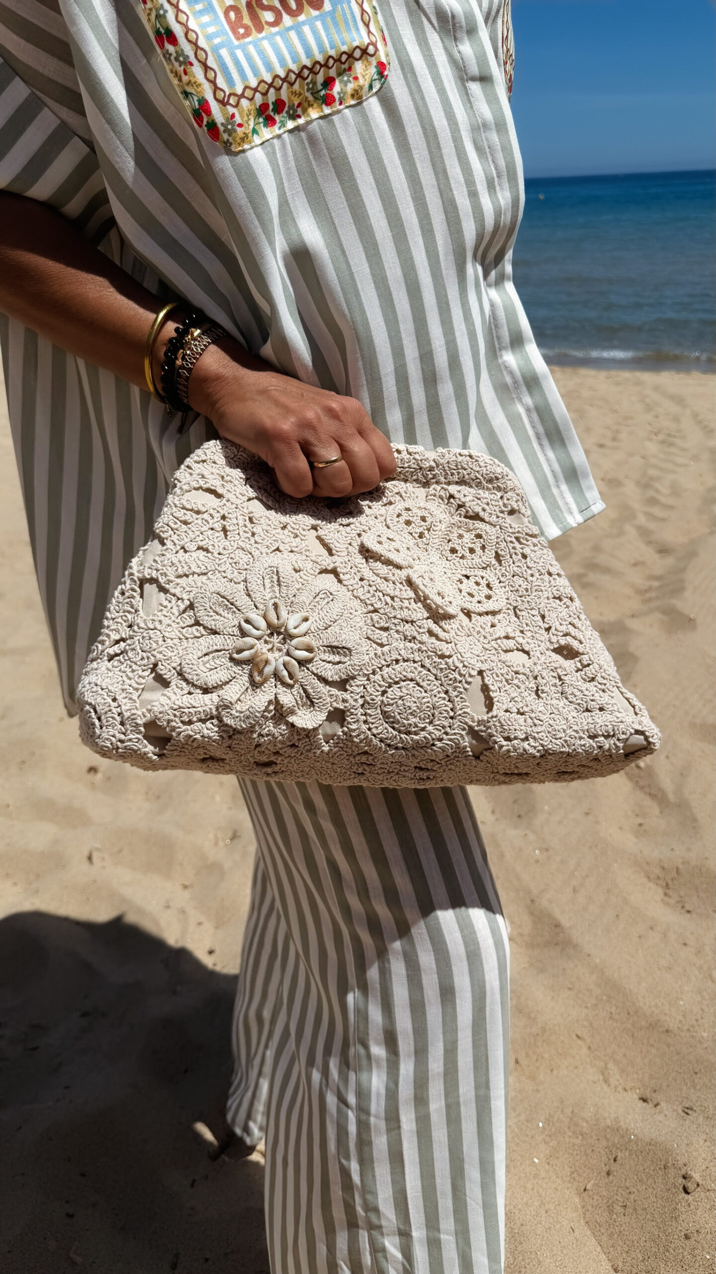 Sac à main femme en crochet beige orné d'une fleur en relief et de coquillages, porté sur une plage en tenue estivale
