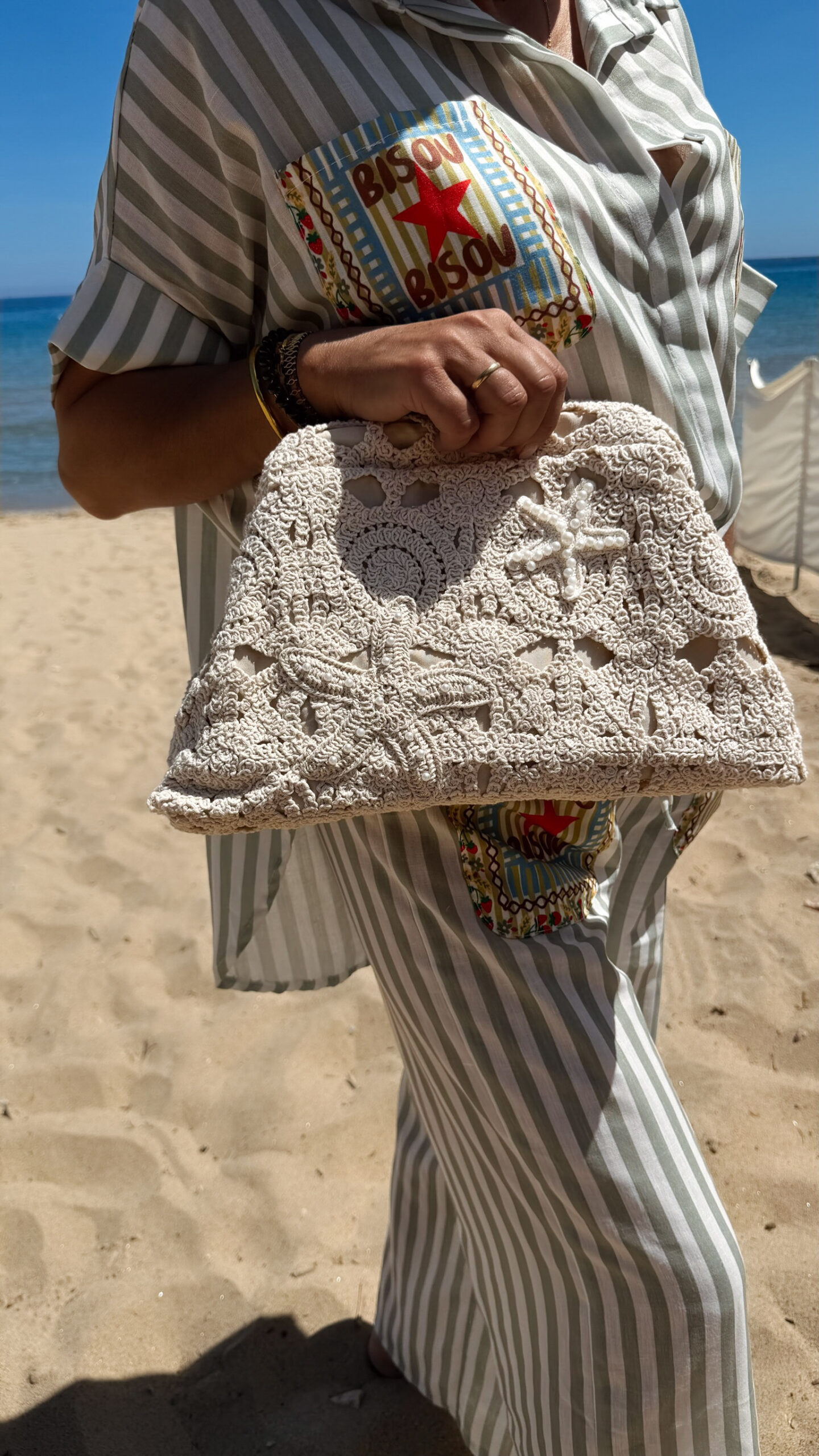 Femme portant un sac à main en crochet beige orné d'étoiles de mer, sur une plage en tenue estivale rayée