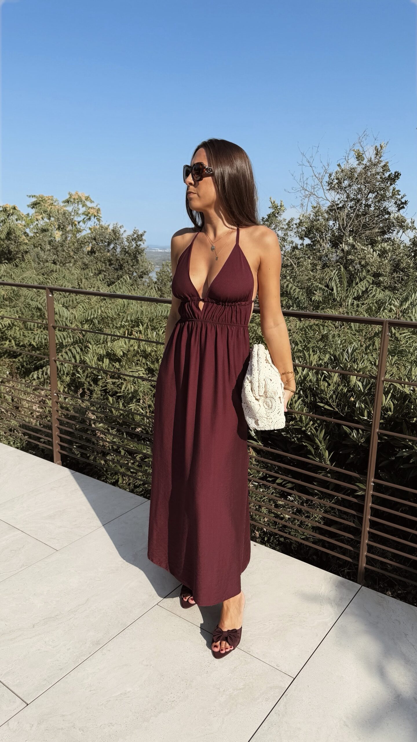 Robe longue dos nu bordeaux à fines bretelles, décolleté plongeant et fente sur la jambe, portée avec des mules à talon assorties.