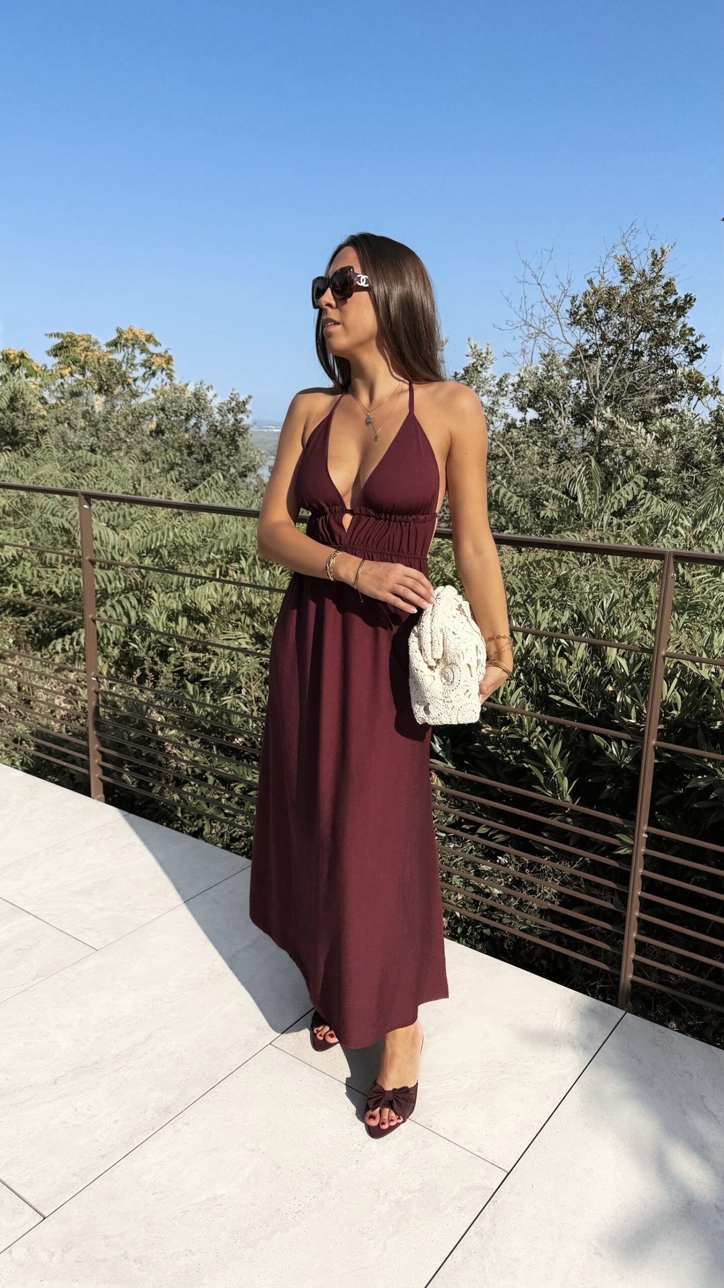 Robe longue dos nu bordeaux à fines bretelles, décolleté plongeant et fente sur la jambe, portée avec des mules à talon assorties.