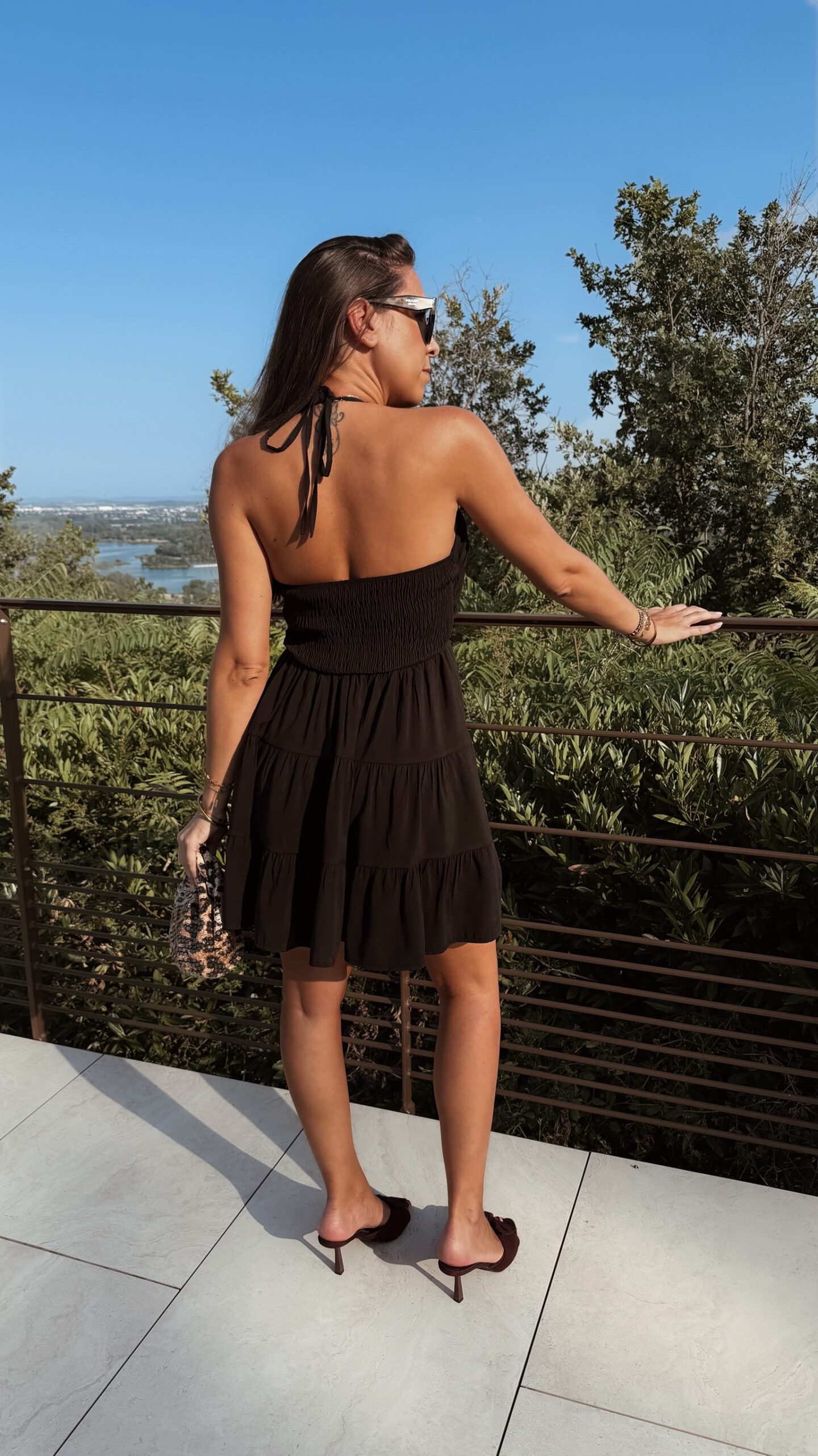 Robe courte noire à fines bretelles avec décolleté drapé, portée avec des mules à talon bordeaux et une pochette imprimé léopard.