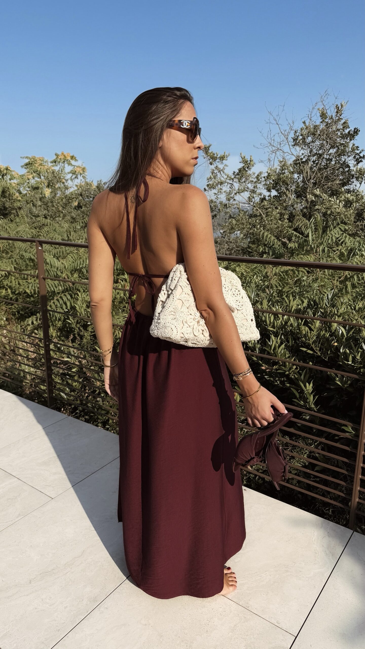 Robe longue dos nu bordeaux à fines bretelles, décolleté plongeant et fente sur la jambe, portée avec des mules à talon assorties.