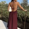 Robe longue dos nu bordeaux à fines bretelles, décolleté plongeant et fente sur la jambe, portée avec des mules à talon assorties.