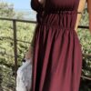 Robe longue dos nu bordeaux à fines bretelles, décolleté plongeant et fente sur la jambe, portée avec des mules à talon assorties.