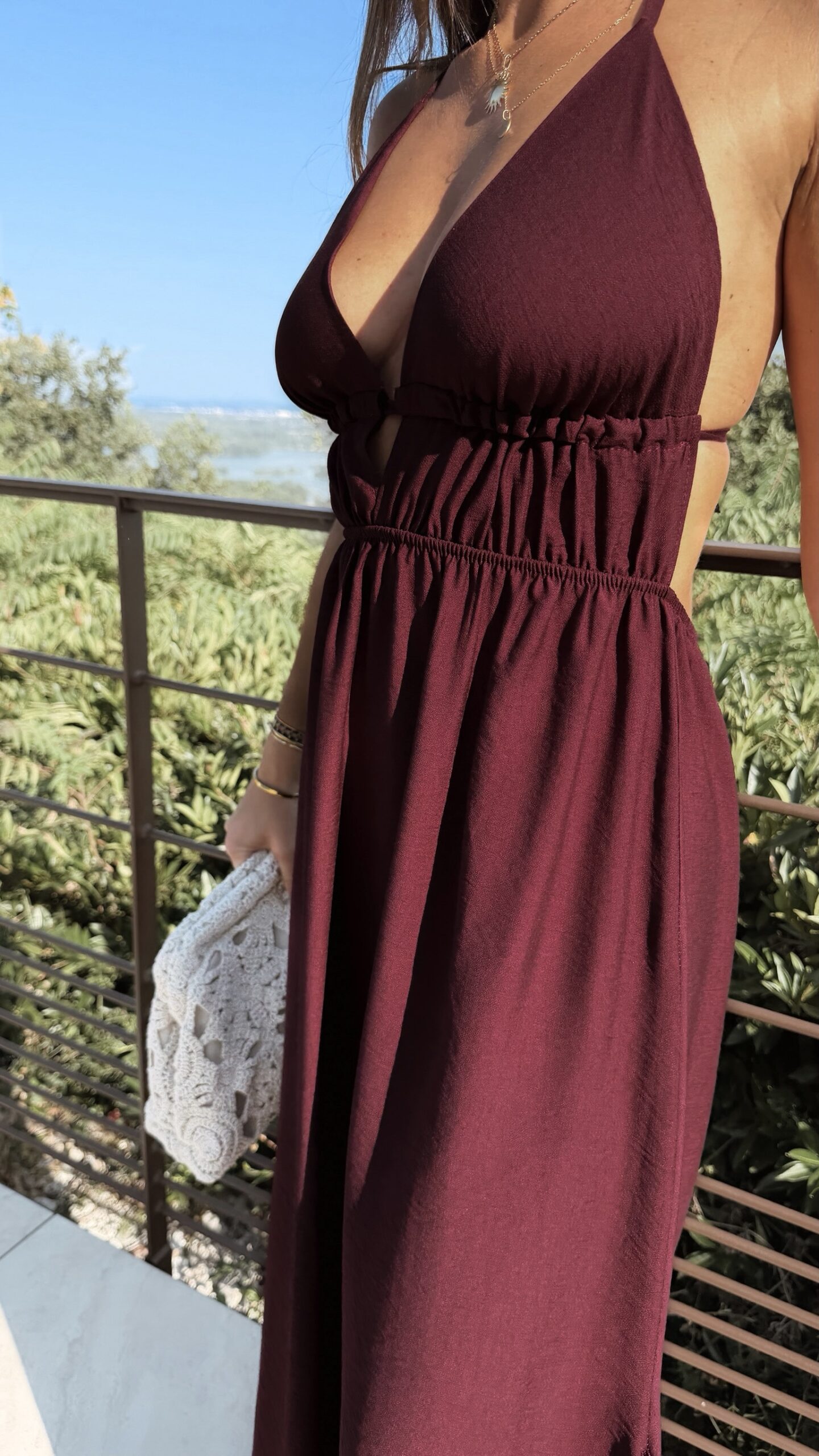 Robe longue dos nu bordeaux à fines bretelles, décolleté plongeant et fente sur la jambe, portée avec des mules à talon assorties.