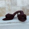 Mules à talon bordeaux en tissu avec nœud sur l'avant, bout pointu et talon fin, portées avec un pantalon assorti.