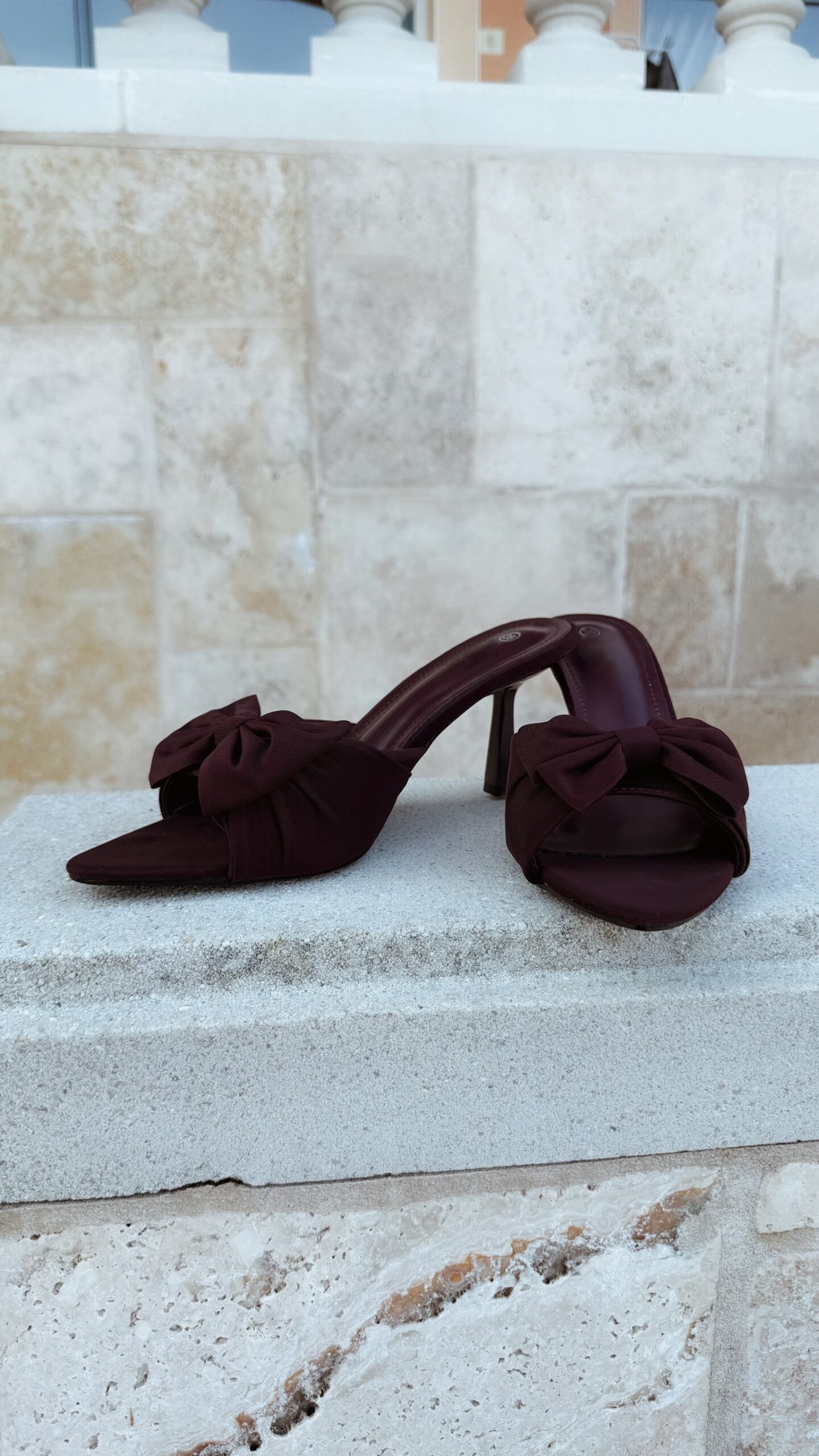 Mules à talon bordeaux en tissu avec nœud sur l'avant, bout pointu et talon fin, portées avec un pantalon assorti.