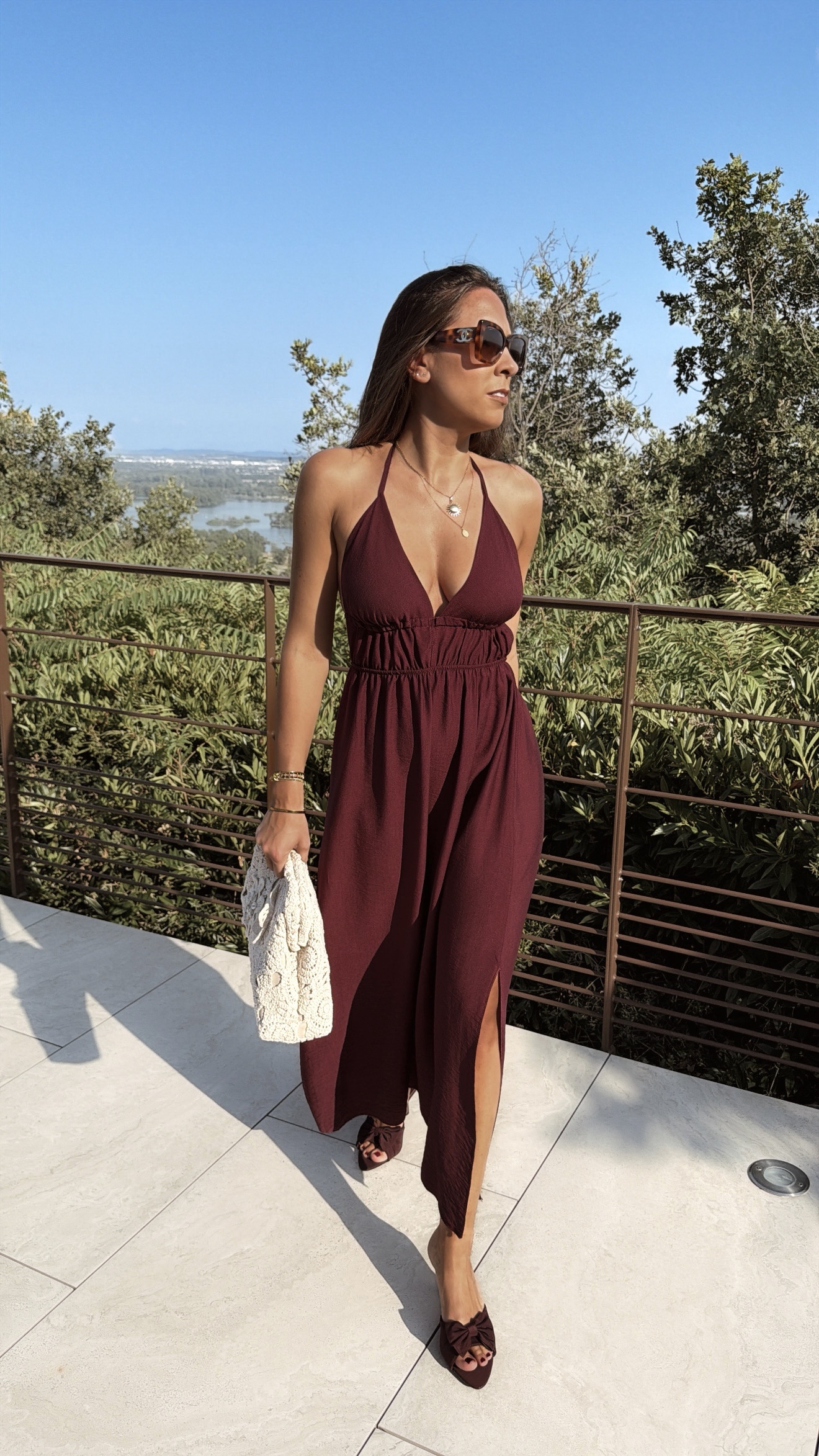 Robe longue dos nu bordeaux à fines bretelles, décolleté plongeant et fente sur la jambe, portée avec des mules à talon assorties.