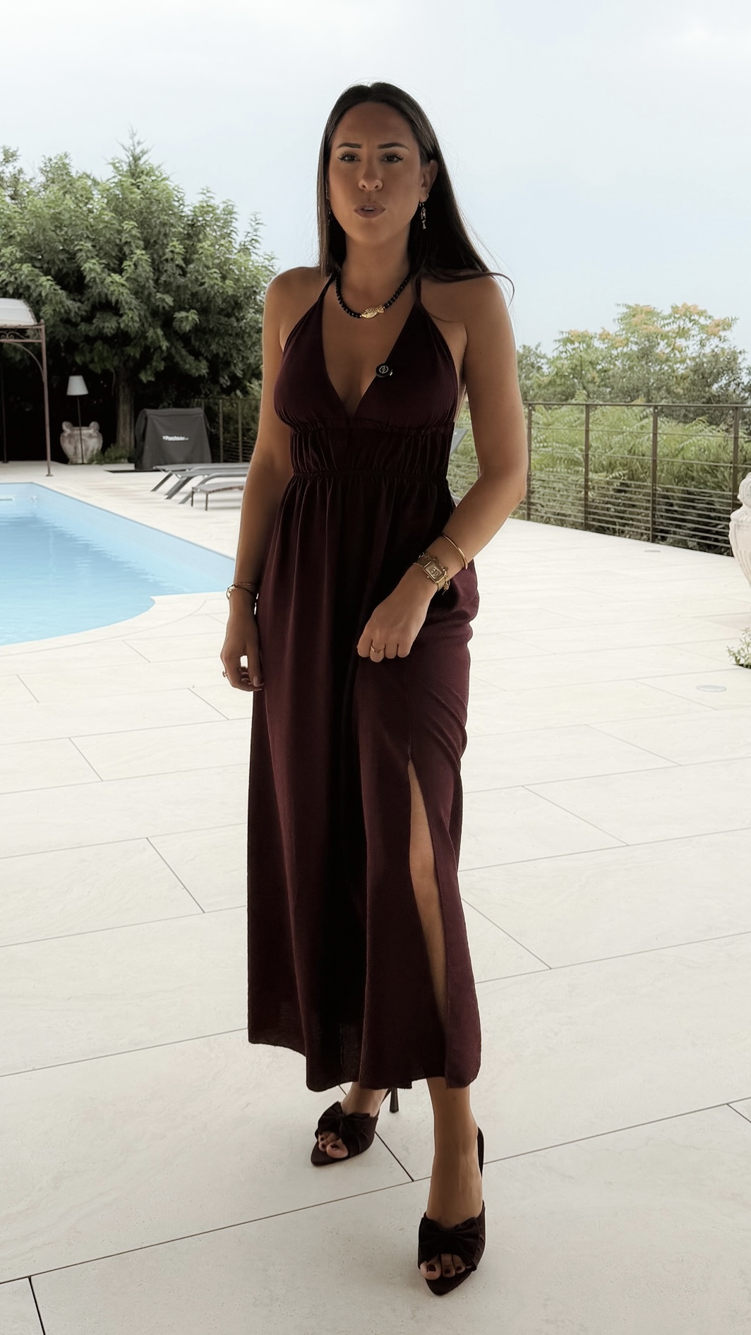 Robe longue dos nu bordeaux à fines bretelles, décolleté plongeant et fente sur la jambe, portée avec des mules à talon assorties.