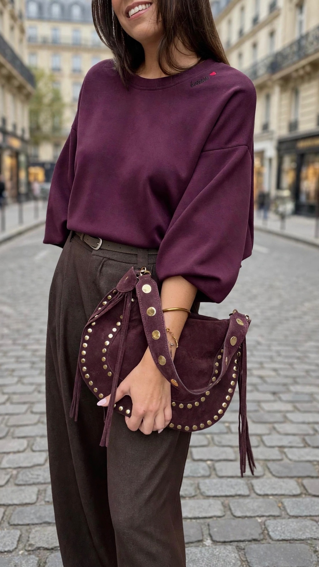 Femme portant un sweatshirt bordeaux oversize, un pantalon large marron et un sac à main bordeaux clouté à détails dorés, pour un look casual chic.