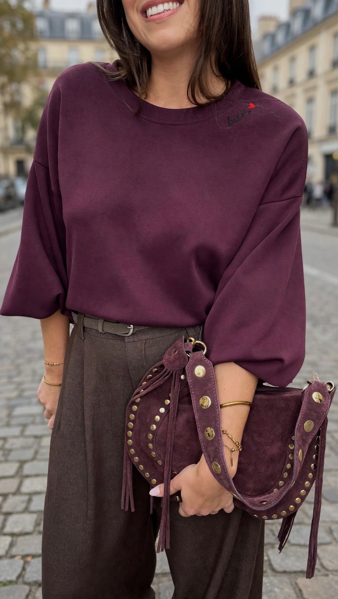 Femme portant un sweatshirt bordeaux oversize, un pantalon large marron et un sac à main bordeaux clouté à détails dorés, pour un look casual chic.