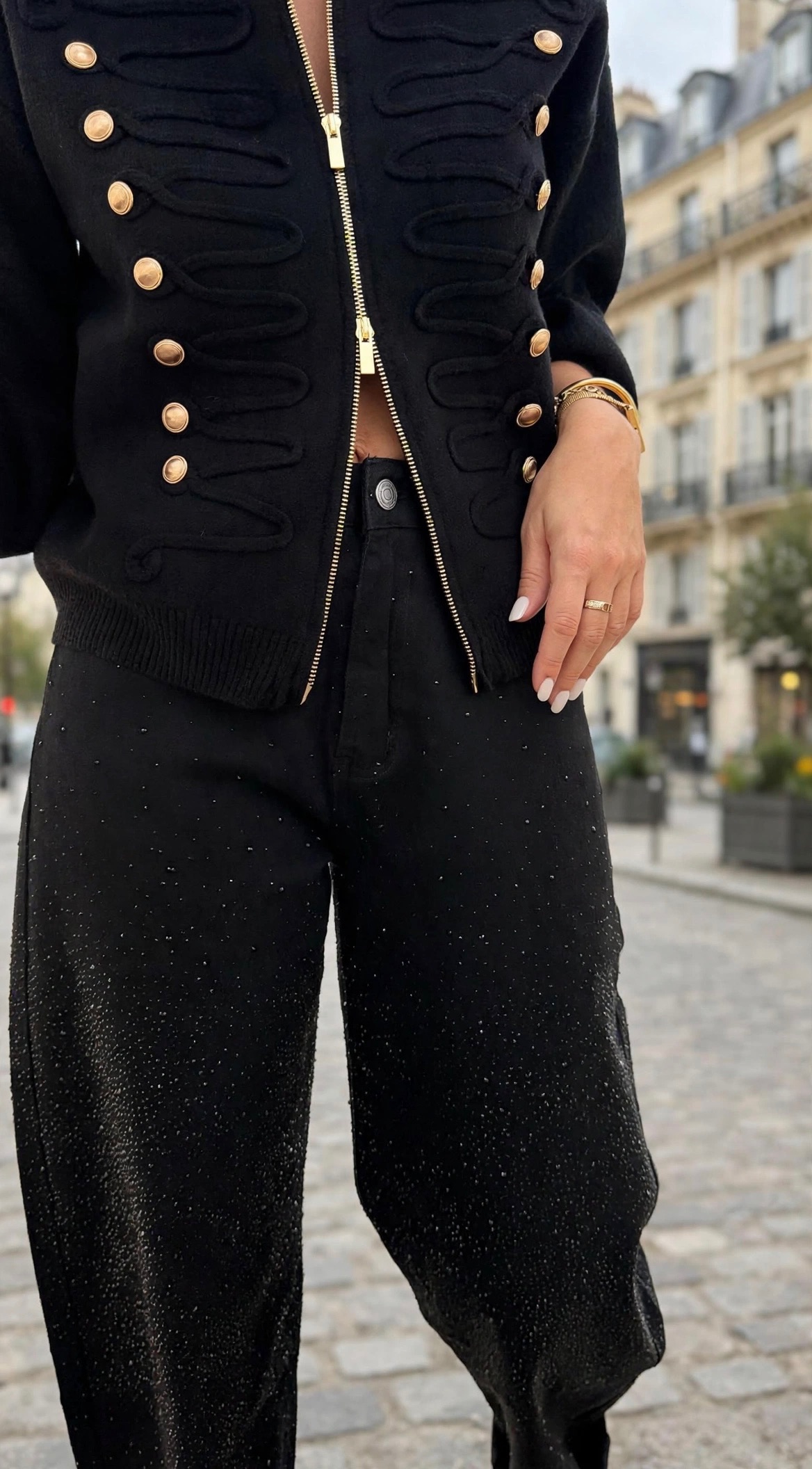 Femme portant une veste noire style officier à boutons dorés, un pantalon noir orné de strass et un sac à main clouté noir, pour un look chic et tendance.