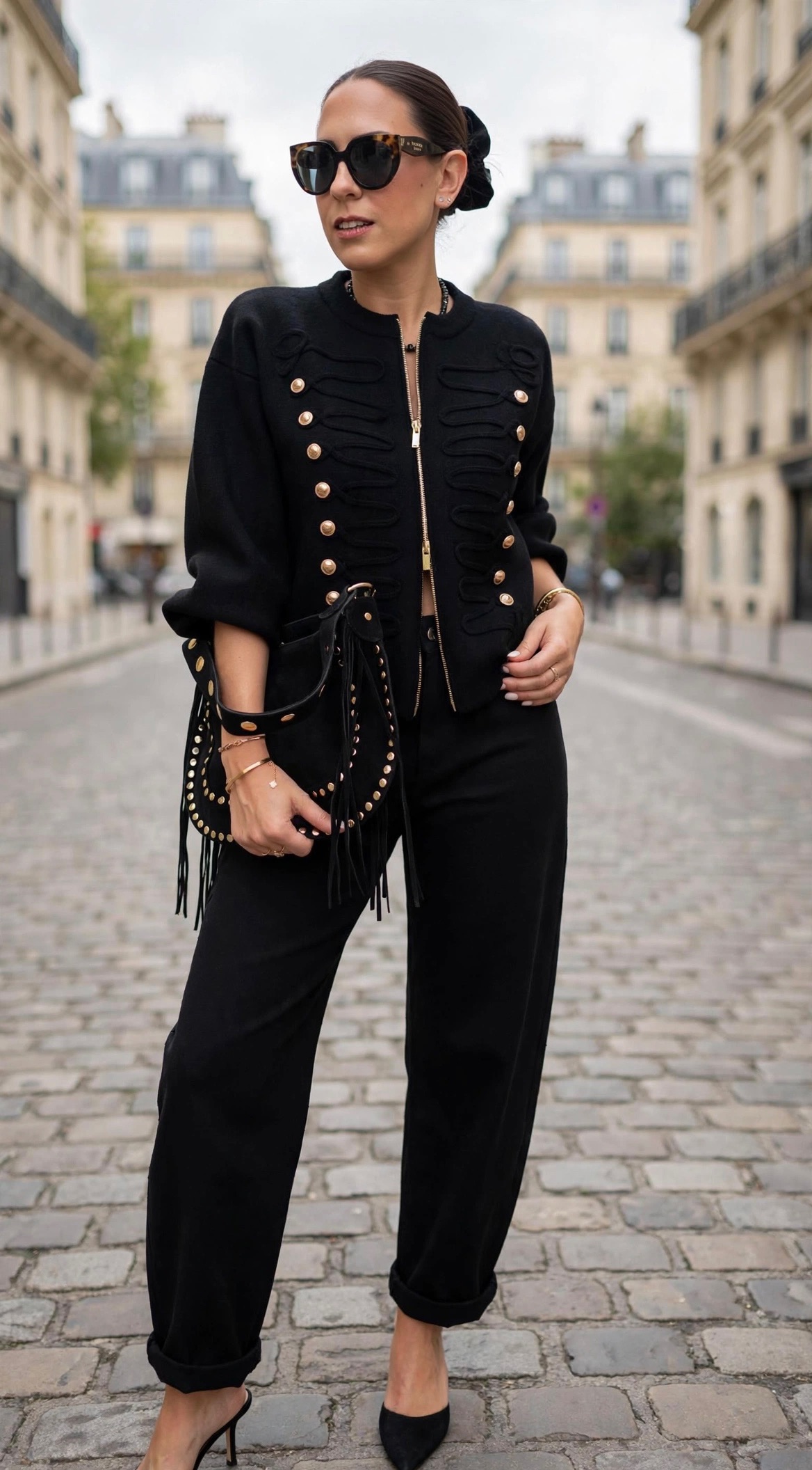 Femme portant une veste noire style officier à boutons dorés, un pantalon noir orné de strass et un sac à main clouté noir, pour un look chic et tendance.