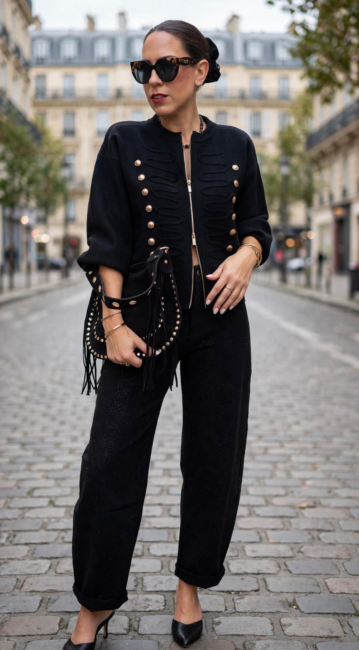 Femme portant une veste noire style officier à boutons dorés, un pantalon noir orné de strass et un sac à main clouté noir, pour un look chic et tendance.